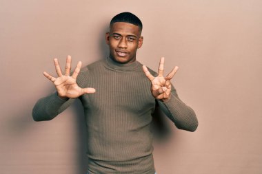 Balıkçı yaka kazak giyen genç siyahi bir adam sekiz numaralı parmaklarını gösterip onu gösterirken kendinden emin ve mutlu bir şekilde gülümsüyor.. 