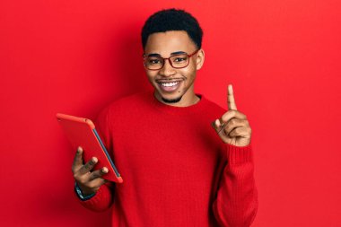 Young african american man using touchpad device surprised with an idea or question pointing finger with happy face, number one 