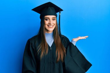 Mezuniyet şapkası ve tören cübbesi giymiş güzel esmer bir kadın neşeli bir şekilde gülümsüyor ve avuç içi kameraya bakıyor.. 