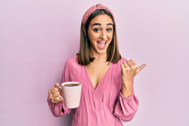 Genç esmer kız bir fincan kahve içiyor başparmağıyla yan tarafa bakıyor ve ağzı açık gülümsüyor. 