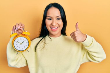 Burnuna piercing takan, elinde çalar saat tutan güzel İspanyol kadın mutlu ve pozitif gülümsüyor, başparmağıyla mükemmel ve onay işareti yapıyor. 