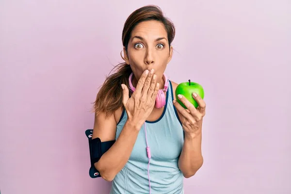 年轻的拉丁女人穿着运动服 用耳机 用手捂着嘴吃着青苹果 既震惊又害怕犯错 惊讶的表情 — 图库照片