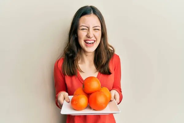 Νεαρή Μελαχρινή Γυναίκα Κρατώντας Πιάτο Φρέσκα Πορτοκάλια Χαμογελώντας Και Γελώντας — Φωτογραφία Αρχείου