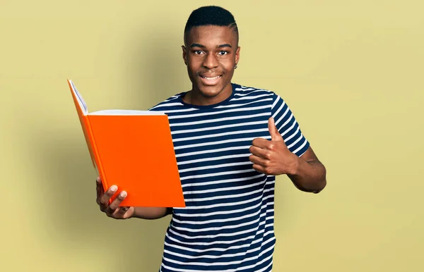 Młody Afroamerykanin Trzyma Książkę Uśmiechnięty Szczęśliwy Pozytywny Kciuk Góry Robi — Zdjęcie stockowe