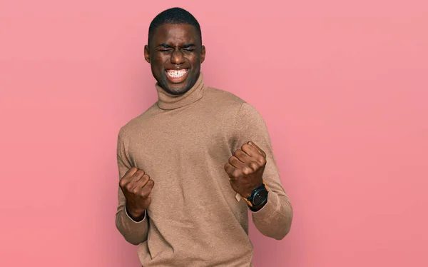 カジュアルな冬のセーターを着ている若いアフリカ系アメリカ人の男性は非常に満足し 腕を上げて勝者のジェスチャーを行う興奮し 笑顔と成功のために叫んでいます お祝いのコンセプト — ストック写真