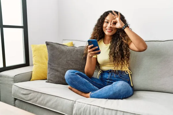 漂亮的惊慌失措的女人坐在家里的沙发上 用智能手机做着一个手势 脸上挂着微笑 脸上挂着幸福的表情 — 图库照片