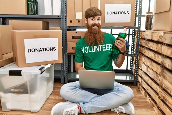 慈善センターでノートパソコンを保持洗剤ボトルを使用して若い赤毛ボランティア男性 — ストック写真