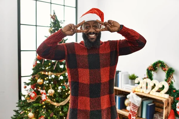 アフリカ系アメリカ人の男はサンタの帽子をかぶってクリスマスツリーのそばに立って顔の上に指で平和のシンボルをやって 明るい勝利を示す笑顔 — ストック写真
