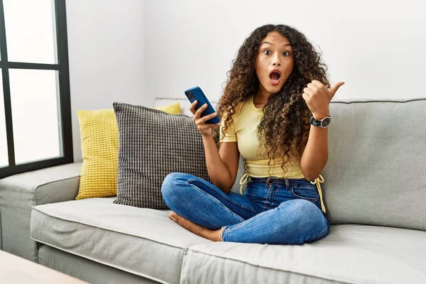 自宅でソファに座っている美しいヒスパニック系の女性は 側に手指で驚いてスマートフォンを使用して 口を開けて驚いて表情 — ストック写真