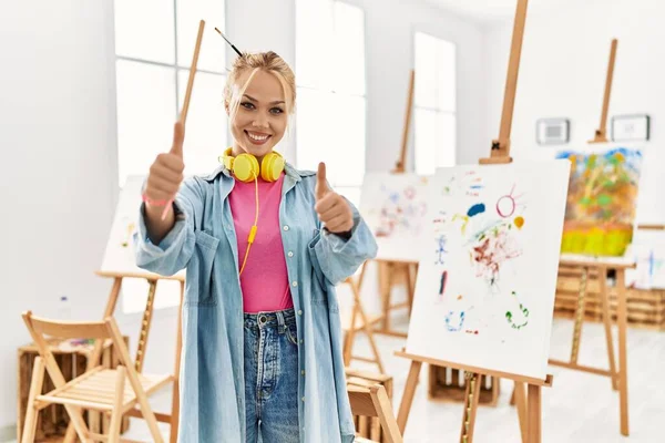 Young caucasian girl at art studio approving doing positive gesture with hand, thumbs up smiling and happy for success. winner gesture.