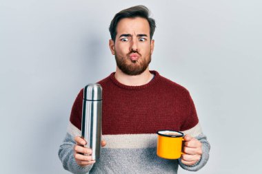 Sakallı yakışıklı beyaz adam bir fincan kahve içiyor, elinde termo ağzıyla balık yüzü yapıyor ve şaşı bakıyor, çılgın ve komik.. 