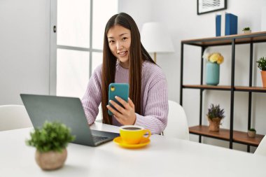 Laptop ve akıllı telefon kullanan Çinli genç bir kız evdeki masada oturuyor..