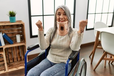 Orta yaşlı, gri saçlı, tekerlekli sandalyede oturan, tasmalı, gururlu, zaferi kutlayan, kollarını kaldırarak heyecanlanan bir kadın. 