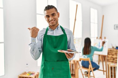 Young latin man standing at art studio pointing thumb up to the side smiling happy with open mouth 