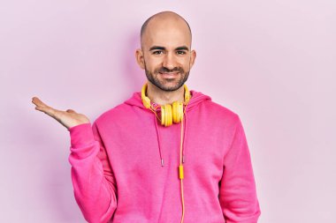 Spor kıyafetleri giyen ve kulaklık takan genç kel bir adam. Gülümseyen, mutlu bir şekilde sunum yapan ve avuç içi kameraya bakan.. 