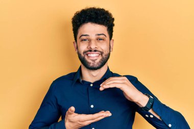 Sakallı, sıradan gömlekli, elleri büyük ve büyük bir işaret gösteren genç bir Arap. Ölçü sembolü. Kameraya bakıp gülümsüyor. ölçüm kavramı. 