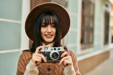 Kışlık şapkalı esmer kadın şehrin dışında klasik kamera kullanarak gülümsüyor.