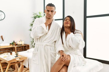 Young latin couple wearing towel standing at beauty center touching painful neck, sore throat for flu, clod and infection 