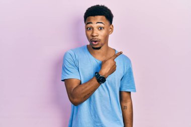 Young african american man wearing casual blue t shirt surprised pointing with finger to the side, open mouth amazed expression. 