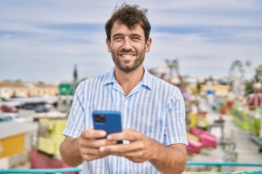 Genç İspanyol adam eğlence parkında akıllı telefon kullanarak mutlu bir şekilde gülümsüyor..