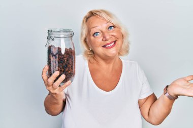 Orta yaşlı sarışın kadın elinde kuru üzüm kasesi tutarak başarıyı mutlu bir gülümsemeyle ve kazanan yüz ifadesiyle kutluyor. 