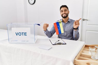 Siyasi seçim kampanyasında sakallı genç yakışıklı bir adam elinde Venezuela bayrağı tutarken mutlu bir şekilde elini ve parmağını yana doğru uzatıyordu. 