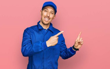 Sakallı, inşaatçı tulumu giyen, gülümseyen ve kameraya bakan kel bir adam iki eliyle ve parmaklarıyla yan tarafı işaret ediyor.. 