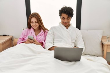 Genç çift yatak odasında dizüstü bilgisayar ve akıllı telefon kullanarak mutlu gülümsüyor..