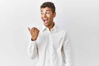Young handsome hispanic man standing over isolated background smiling with happy face looking and pointing to the side with thumb up. 