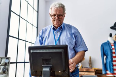 Orta yaşlı, gri saçlı, gülümseyen, kendine güvenen bir adam giyim mağazasında çalışıyor.