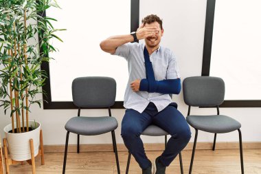 Yakışıklı genç adam, kolunda sakatlıkla bekleme odasında oturuyor. Gülümsüyor ve elleri yüzünde, şaşırtmak için gözlerini kapatıyor. kör kavram. 