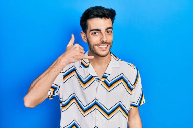 Günlük kıyafetler giymiş, gülümseyen genç İspanyol adam telefonla el ve parmak hareketleri yapıyor telefonda konuşur gibi. iletişim kavramları. 