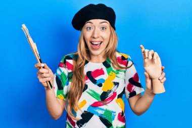 Beyaz kadın sanatçı elinde küçük ahşap manikinler ve boya fırçaları tutarak çılgınlığı kutluyor ve başarıyı hayretle haykırıyor.. 