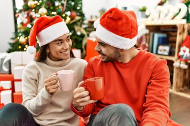 Mutlu gülümseyen ve kahve içen genç İspanyol çift evdeki Noel ağacının yanında yerde oturuyor..