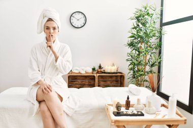 Young blonde woman wearing bathrobe at wellbeing spa asking to be quiet with finger on lips. silence and secret concept. 