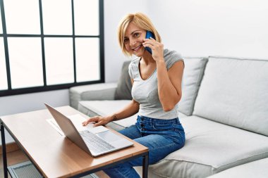 Orta yaşlı sarışın kadın evde dizüstü bilgisayar kullanarak akıllı telefondan konuşuyor.