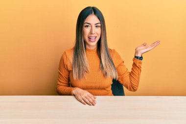 Genç İspanyol kadın, gündelik giysiler içinde masaya oturmuş neşeli bir şekilde gülümsüyor ve avuç içiyle kameraya bakıyor.. 