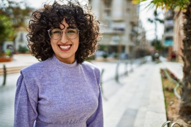 Orta Doğulu genç bir kadın gülümsüyor. Sokakta gözlük takıyor.