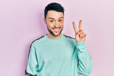 Gündelik kıyafetler giyen, gülümseyen ve kameraya göz kırpan genç İspanyol adam zafer işareti yapıyor. İki numara.. 