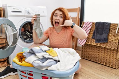 Kirli çamaşırlarını çamaşır makinesine koyup gülümseyen, el ve parmaklarıyla telefonla konuşan genç, kızıl saçlı bir kadın. iletişim kavramları. 
