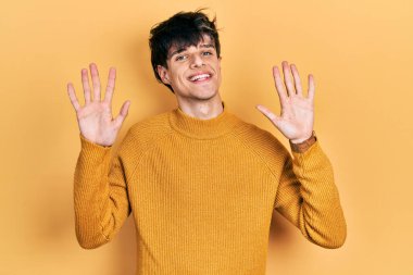 Yakışıklı hippi genç adam gündelik sarı süveter giyiyor ve parmaklarıyla on numarayı gösterip gülümsüyor kendinden emin ve mutlu.. 