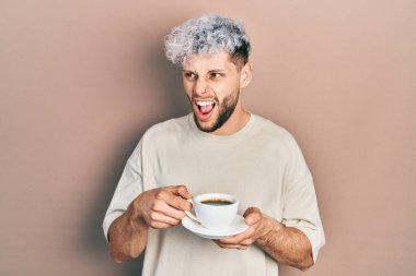 Modern boyalı saçlı genç İspanyol bir adam bir fincan kahve içiyor kızgın ve kızgın çığlıklar atıyor sinirlenmiş ve öfkeyle bağırıyor. öfke ve agresif konsept. 