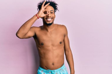 Sakallı, mayo giyen, el hareketleriyle gülümseyen, mutlu yüzüyle parmaklarının arasından bakan Afro-Amerikan genç adam.. 