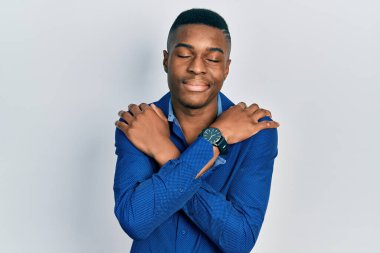 Genç, Afro-Amerikan bir adam günlük kıyafetler giyip kendine sarılıyor, mutlu ve pozitif, kendinden emin gülümsüyor. Sevgi ve özgüven. 