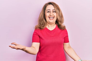 Orta yaşlı beyaz kadın, günlük kıyafetler giyip neşeli bir şekilde kollarını açıp gülümsüyor. Hoş geldin, olumlu ve kendinden emin selamlar. 