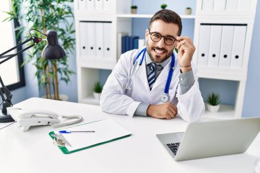 Young hispanic man wearing doctor uniform using laptop sitting at clinic