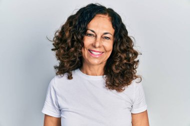 Orta yaşlı İspanyol kadın, mutlu ve havalı bir gülümsemesi olan günlük beyaz tişört giyiyor. şanslı kişi. 