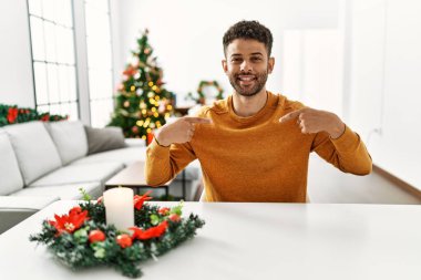 Arap genç adam Noel ağacının yanında masada oturuyor. Yüzünde gülümsemeyle kendinden emin görünüyor. Kendini gururlu ve mutlu bir şekilde gösteriyor.. 