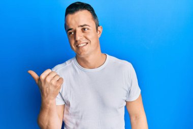 Handsome young man wearing casual white t shirt pointing thumb up to the side smiling happy with open mouth 