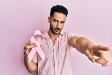 Elinde pembe kanser kurdelesi olan genç İspanyol bir adam kamerayı işaret ediyor ve sen kendine güvenen ciddi bir hareket yapıyorsun. 